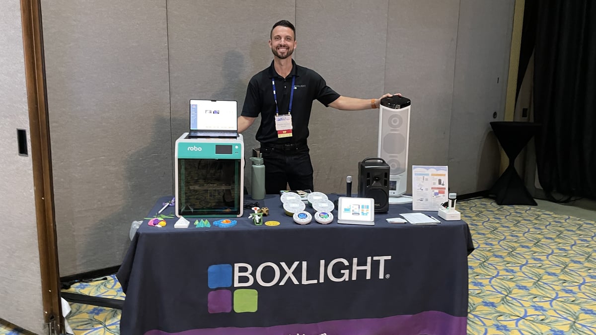 Braydon Moreno, STEM Director at Boxlight, standing behind a table covered with Boxlight solutions including the award-winning Robo E4 3D printer which comes bundled with MyStemKits curriculum, a set of Labdisc all-in-one-science labs, a FrontRow Lyrik portable audio system, a FrontRow Juno audio speaker tower, and the Action! teacher mic. 